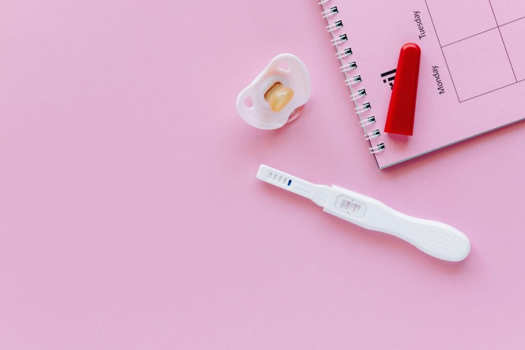 A positive pregnancy test on a pink surface with a pacifier and planner.