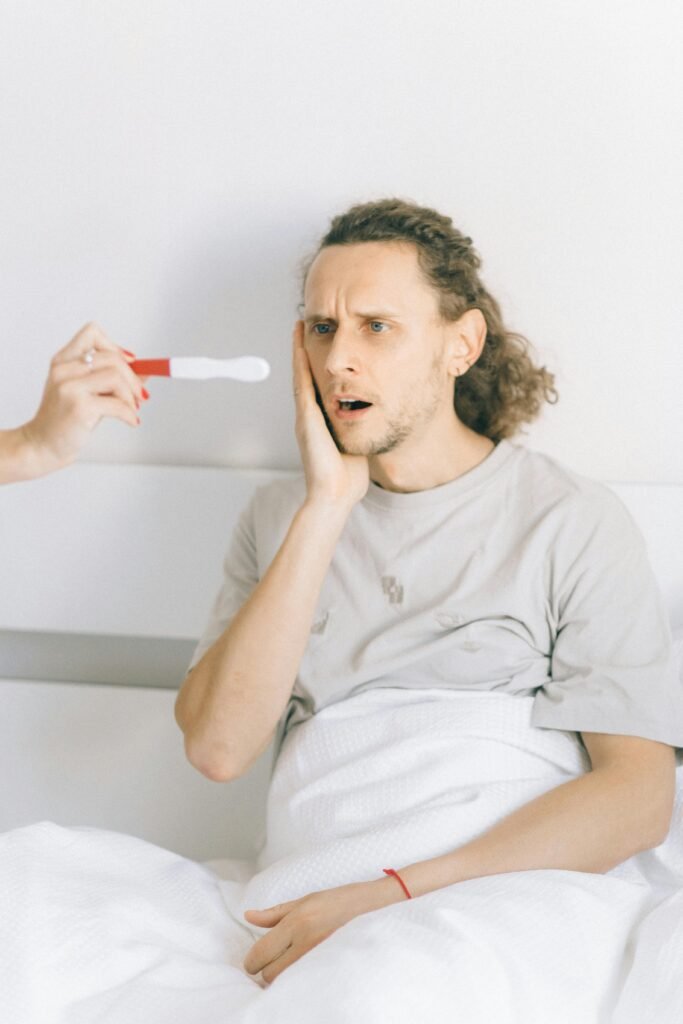 A man in a bedroom shows a surprised expression as a pregnancy test is shown to him.