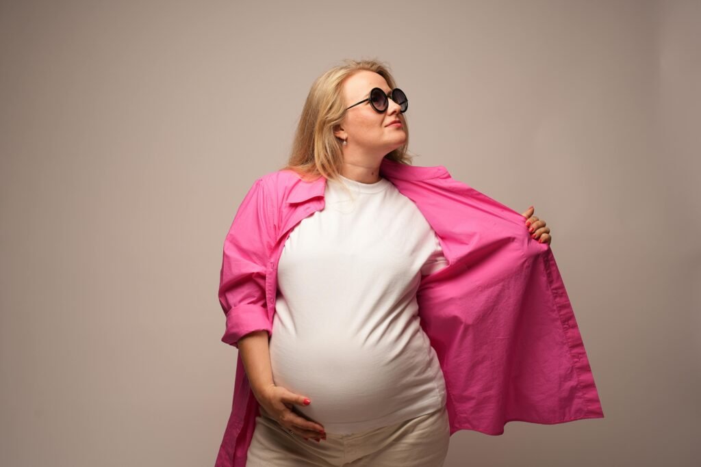 Confident pregnant woman posing stylishly in pink jacket and sunglasses during a studio shoot.