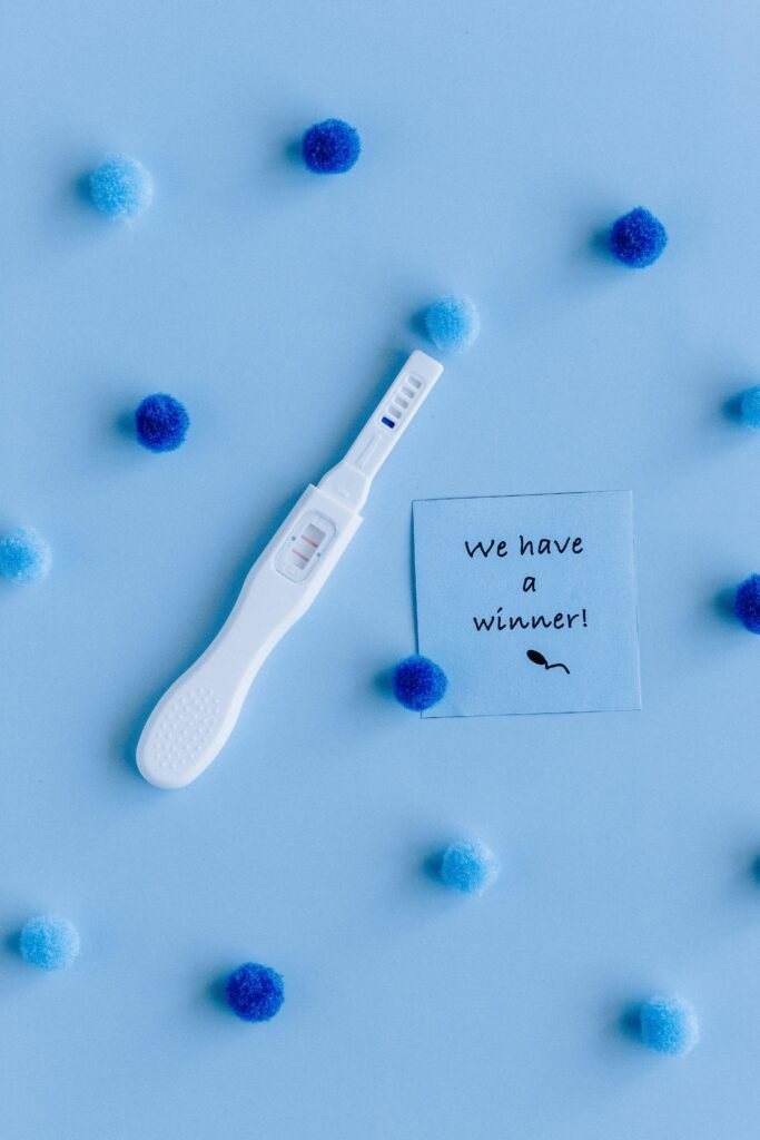 Positive pregnancy test surrounded by pom-poms on blue background with 'We have a winner!' note.