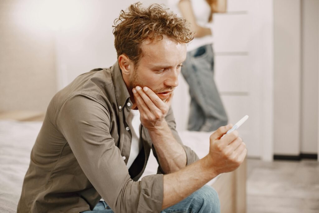 A concerned man sitting on a bed, holding a pregnancy test, deep in thought.
